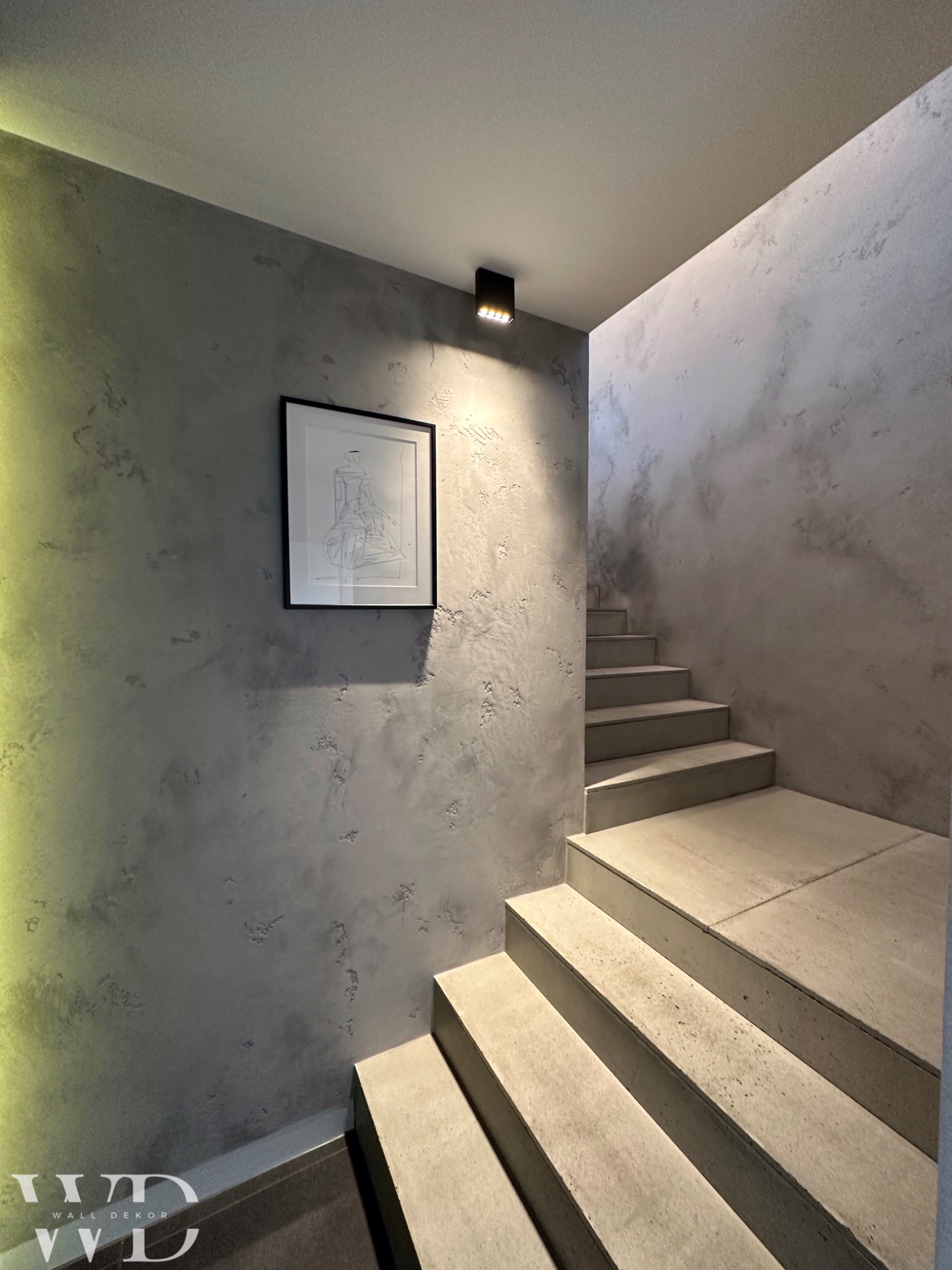 Klatka schodowa z betonowymi schodami i ścianą pokrytą szarą, dekoracyjną fakturą z widocznym rysunkiem w czarnej ramie i nowoczesnym oświetleniem.