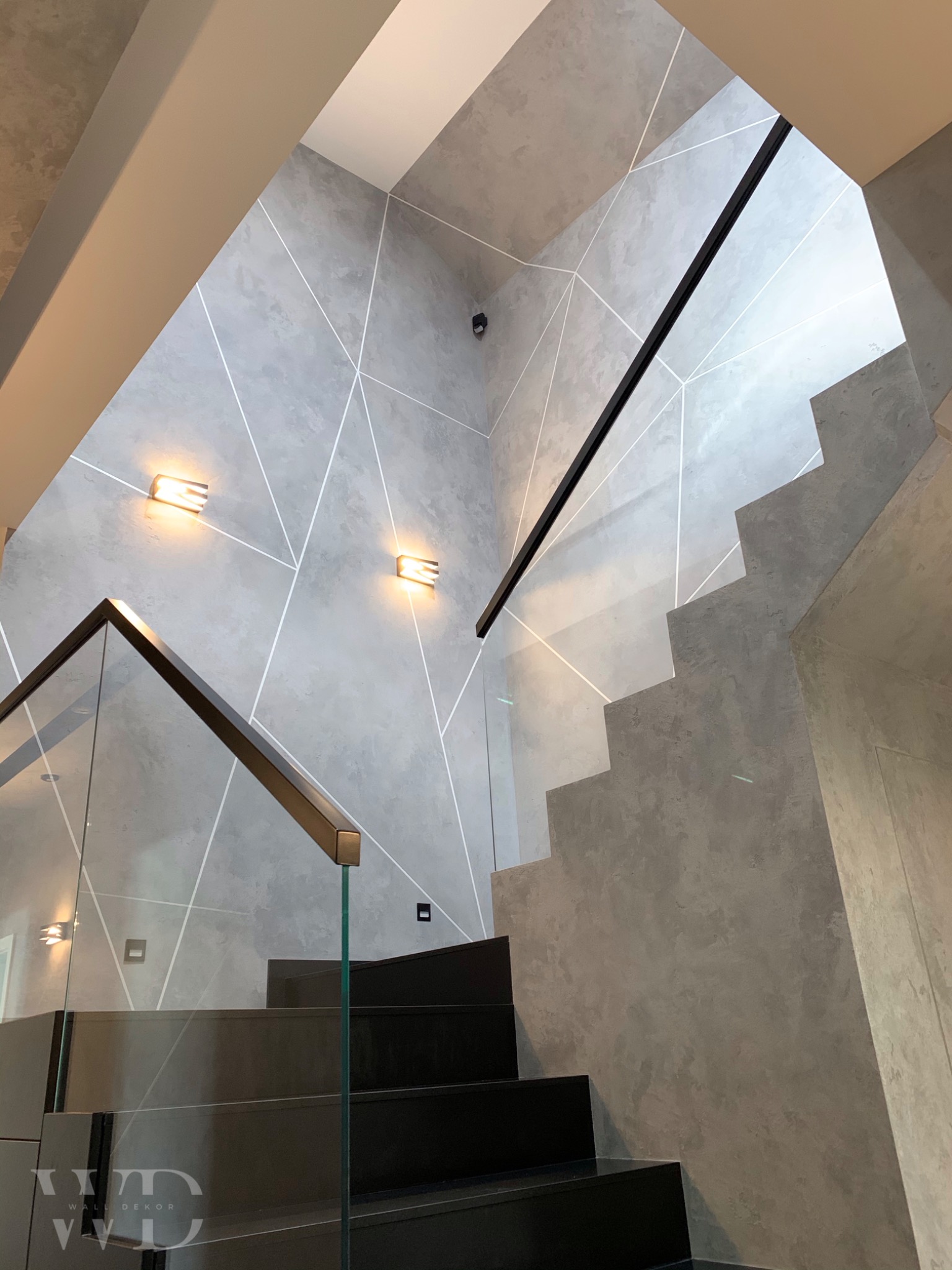 Nowoczesne schody z czarnymi stopniami i szklaną balustradą na tle szarej ściany z geometrycznym wzorem i oświetleniem punktowym.