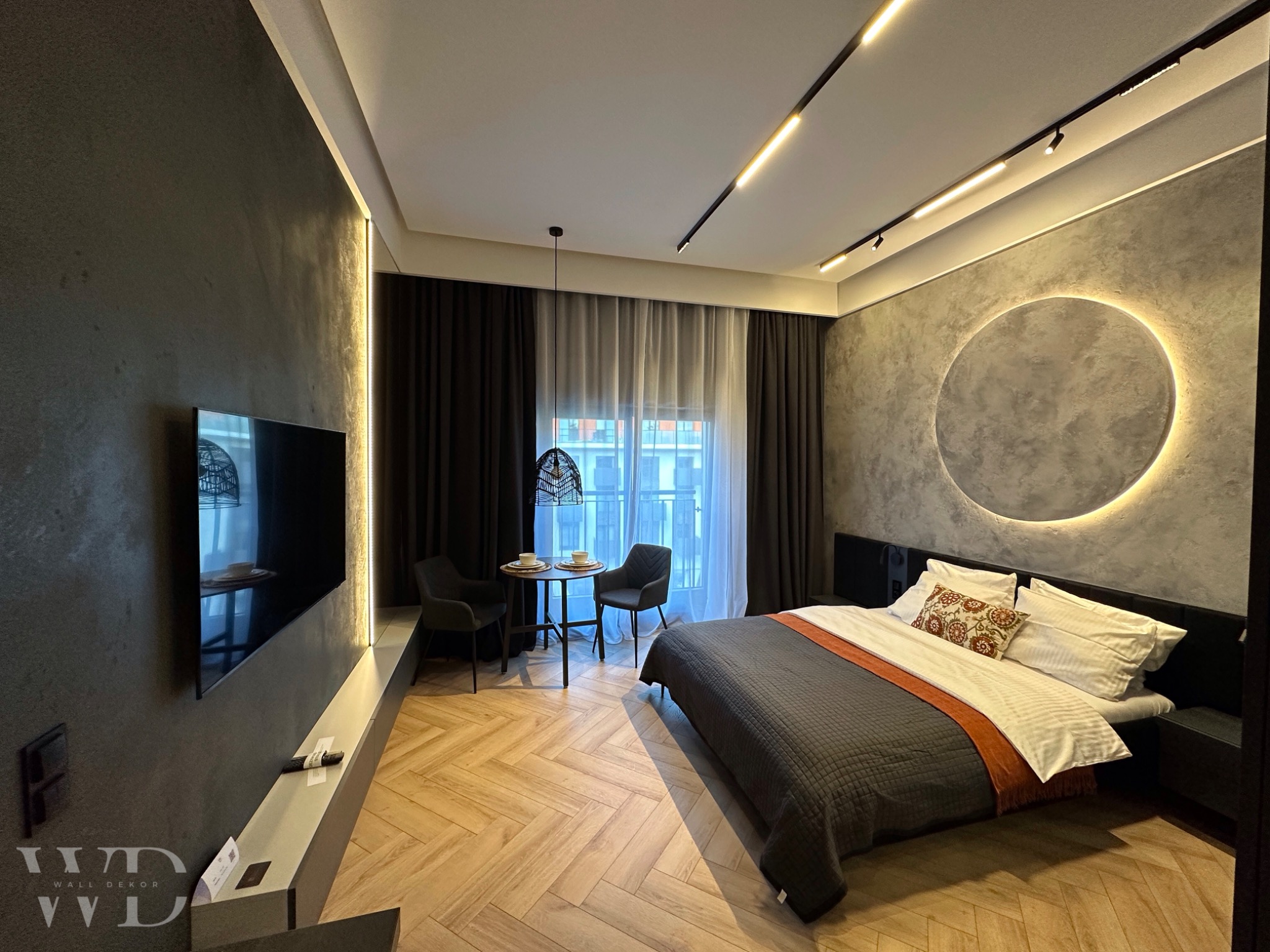 Sypialnia z szarą ścianą z efektem betonu i okrągłą dekoracją podświetloną LED, parkiet w jodełkę, ciemne zasłony i minimalistyczny zestaw stolik z krzesłami przy oknie.