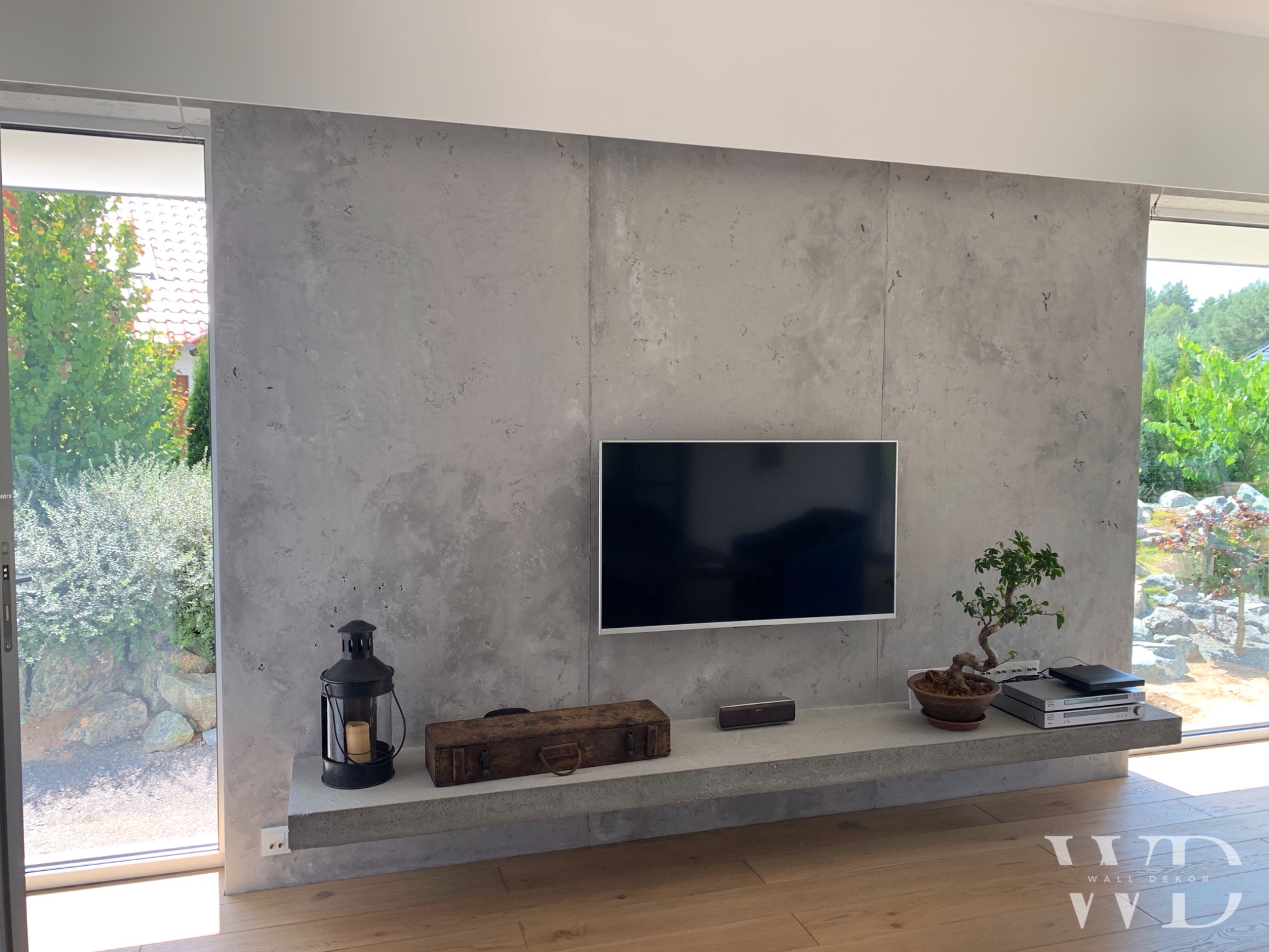 Ściana w salonie wykończona szarym betonem architektonicznym, z wiszącą półką, telewizorem i dekoracjami, widok na zieleń z okna.