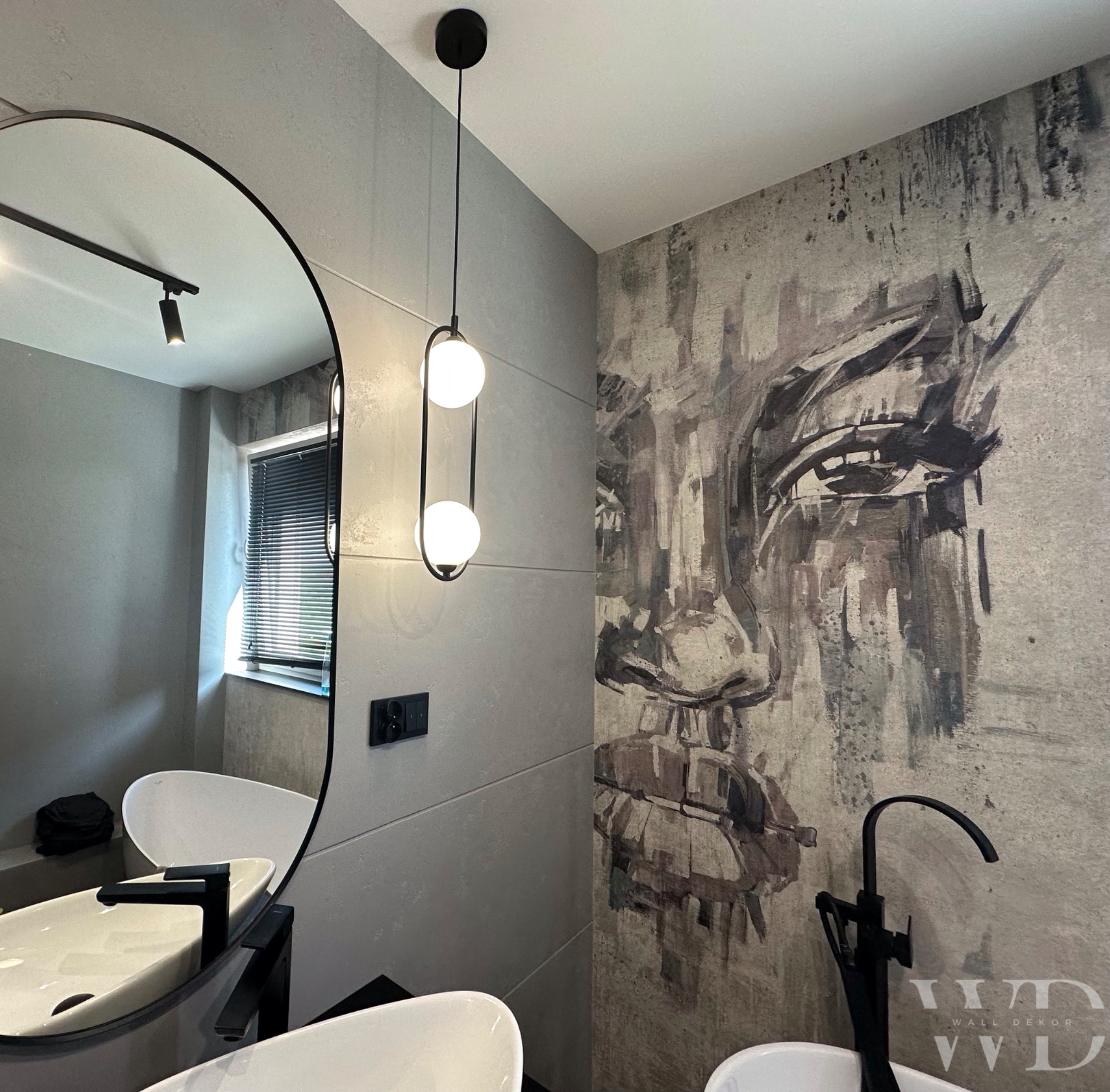 Nowoczesna łazienka z szarą ścianą ozdobioną abstrakcyjnym portretem, okrągłe lustro odbija białą wannę i umywalkę, czarna armatura i lampa z kulistymi kloszami.