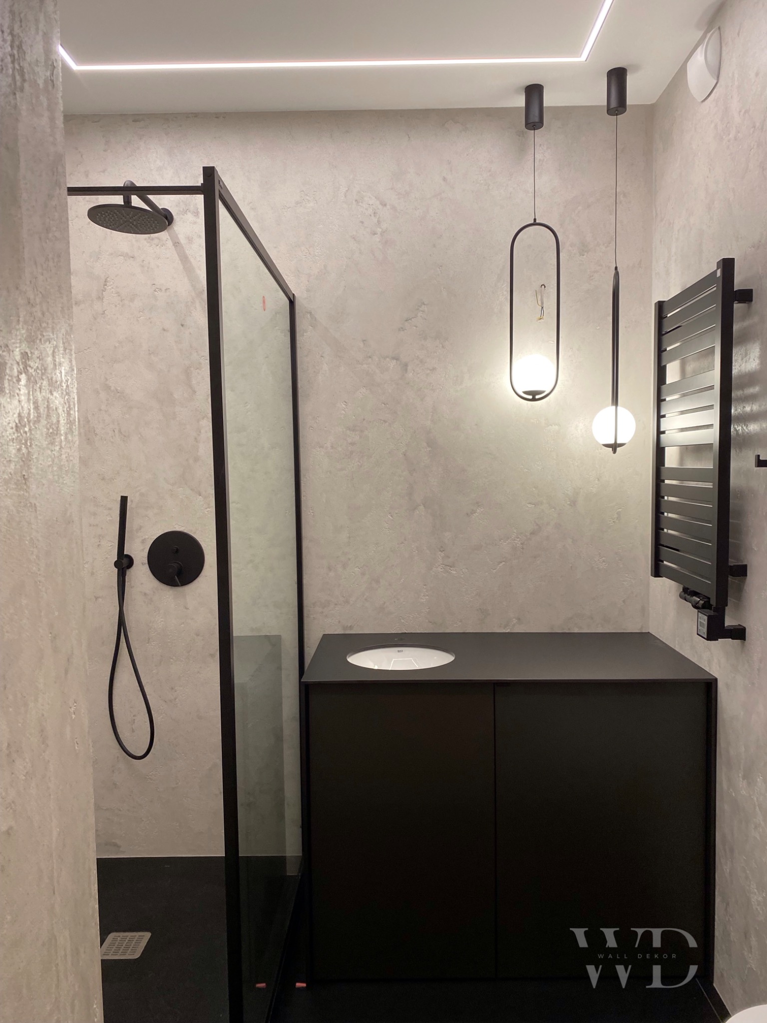 Nowoczesna łazienka z kabiną prysznicową typu walk-in, czarną armaturą, szafką pod umywalką i grzejnikiem drabinkowym na ścianie pokrytej dekoracyjnym tynkiem.