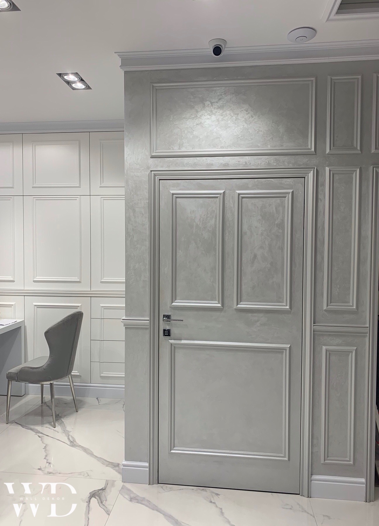 Eleganckie szare drzwi wewnętrzne z panelami dekoracyjnymi na ścianie, wykończone w stylu klasycznym, z widoczną fakturą tynku dekoracyjnego i marmurową posadzką.