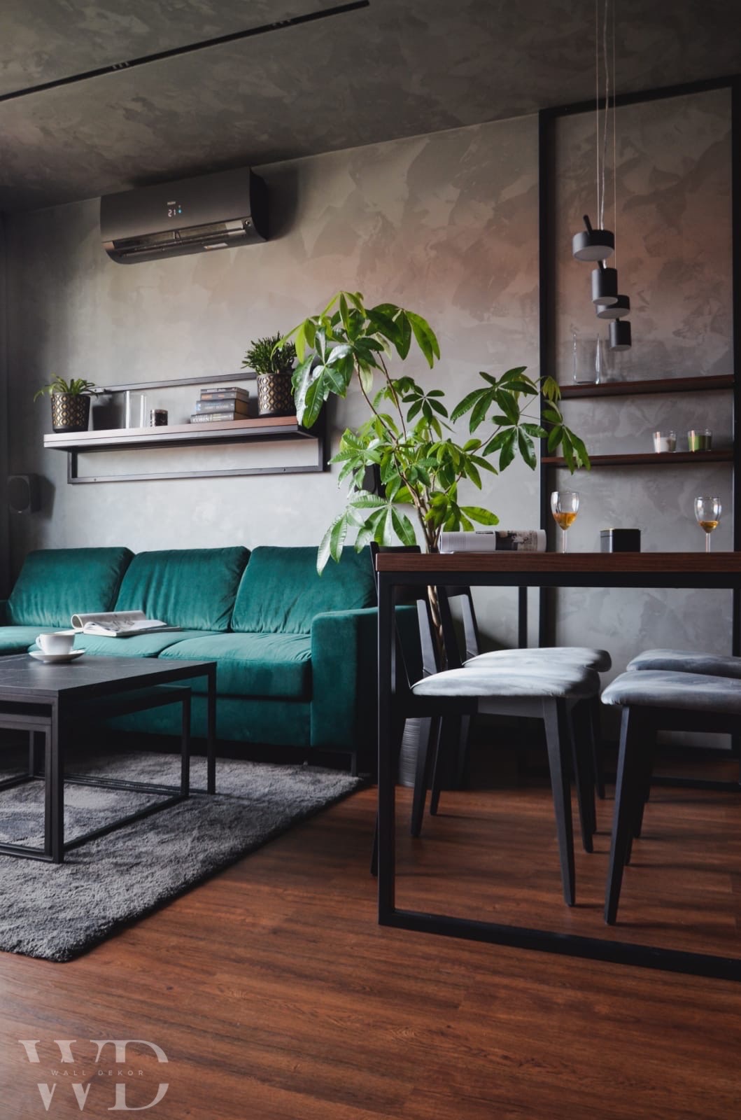 Nowoczesne wnętrze z szarą ścianą o fakturze betonu, zieloną sofą i drewnianą podłogą, akcentowane roślinami doniczkowymi i czarnymi meblami.