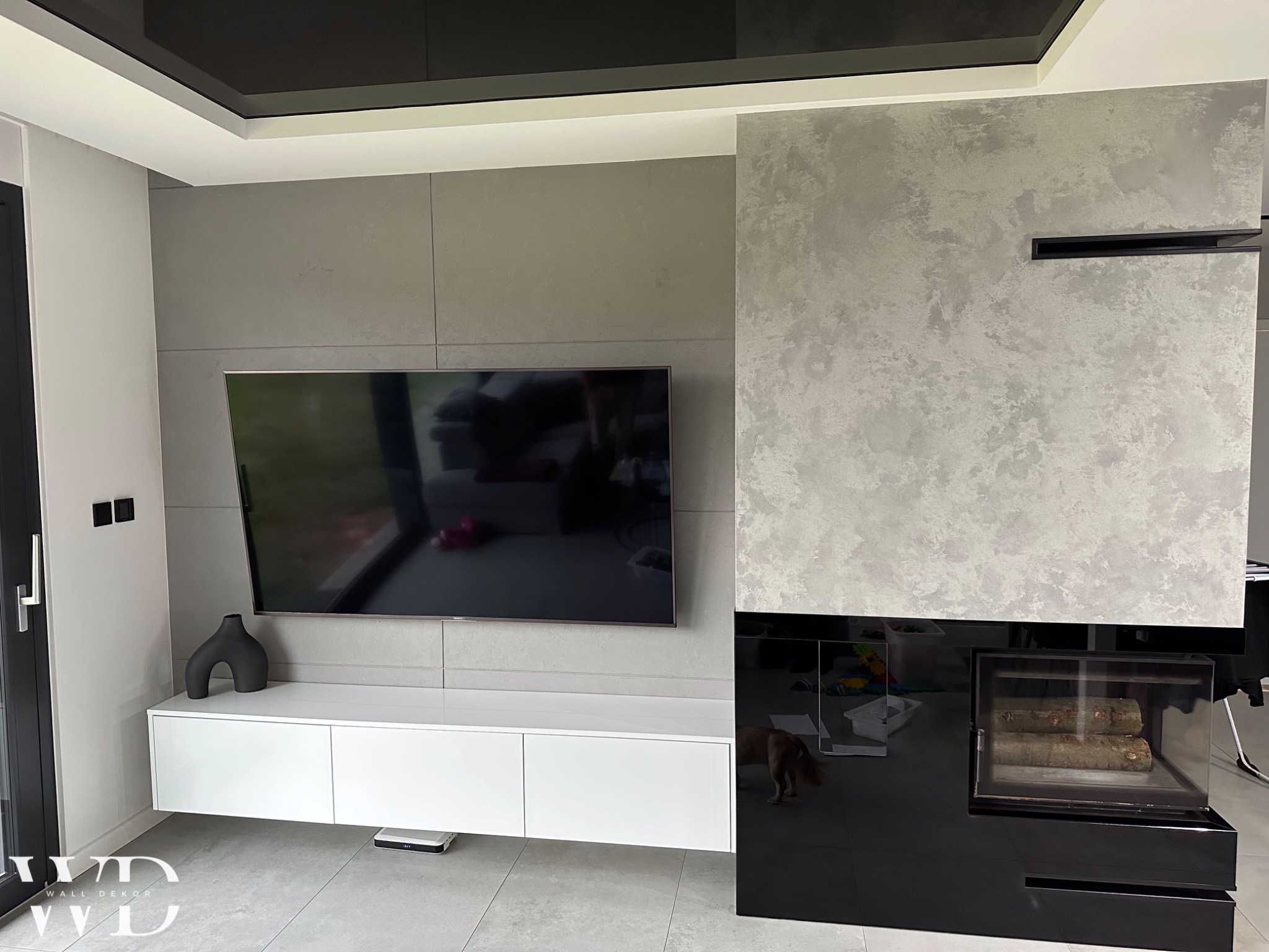 Nowoczesny salon z szarą ścianą z efektem betonu, podwieszanym telewizorem, białymi szafkami RTV i kominkiem z czarnym szkłem.