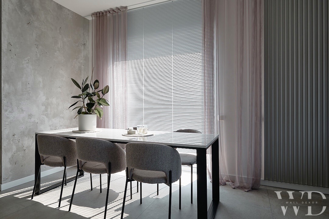 Jadalnia z betonową ścianą o nieregularnej fakturze, minimalistycznym stołem z białym blatem i szarymi krzesłami, doświetlona przez żaluzje i delikatne, różowe zasłony.