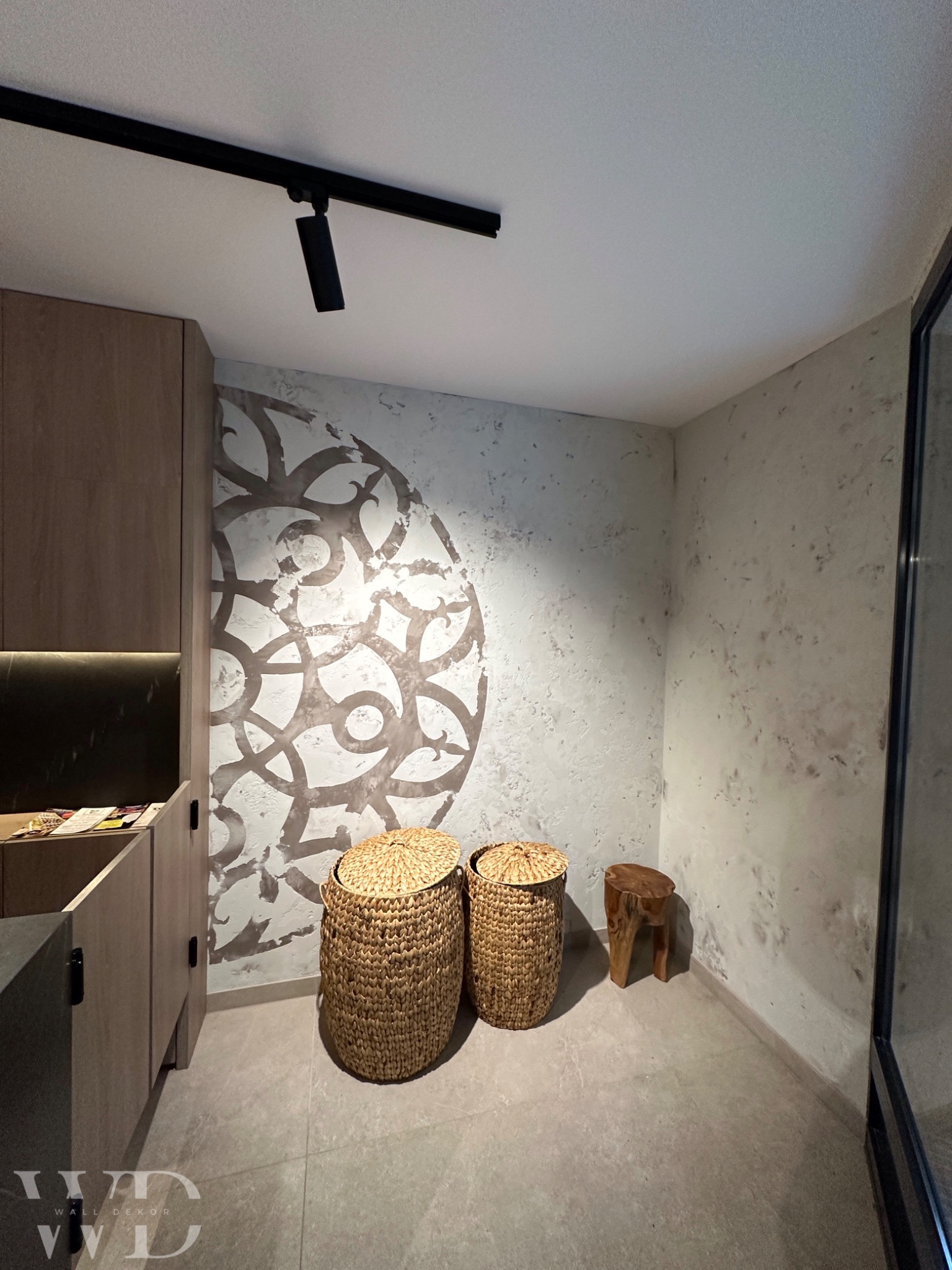 Ściana z dekoracyjnym tynkiem imitującym ornament, uzupełniona o wiklinowe kosze i stołek z pnia drzewa, oświetlona punktowo.