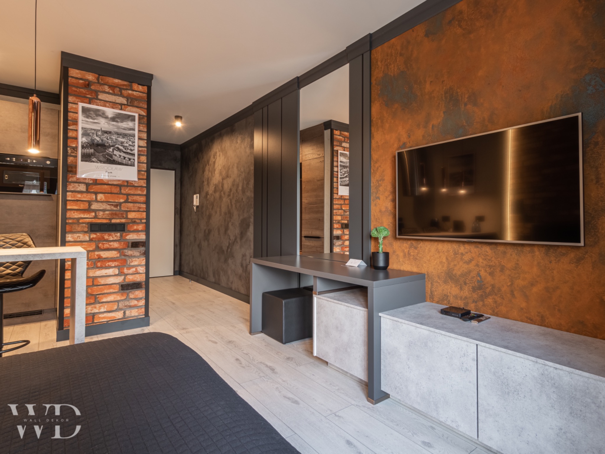 Nowoczesne wnętrze mieszkania z dekoracyjną ścianą w odcieniach rdzy, ceglanym fragmentem ściany z plakatem, szarą zabudową meblową i jasną podłogą z paneli.