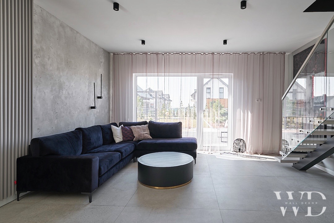 Nowoczesny salon z ciemnoniebieską narożną sofą, okrągłym stolikiem kawowym, ścianą z efektem betonu i schodami ze szklaną balustradą.