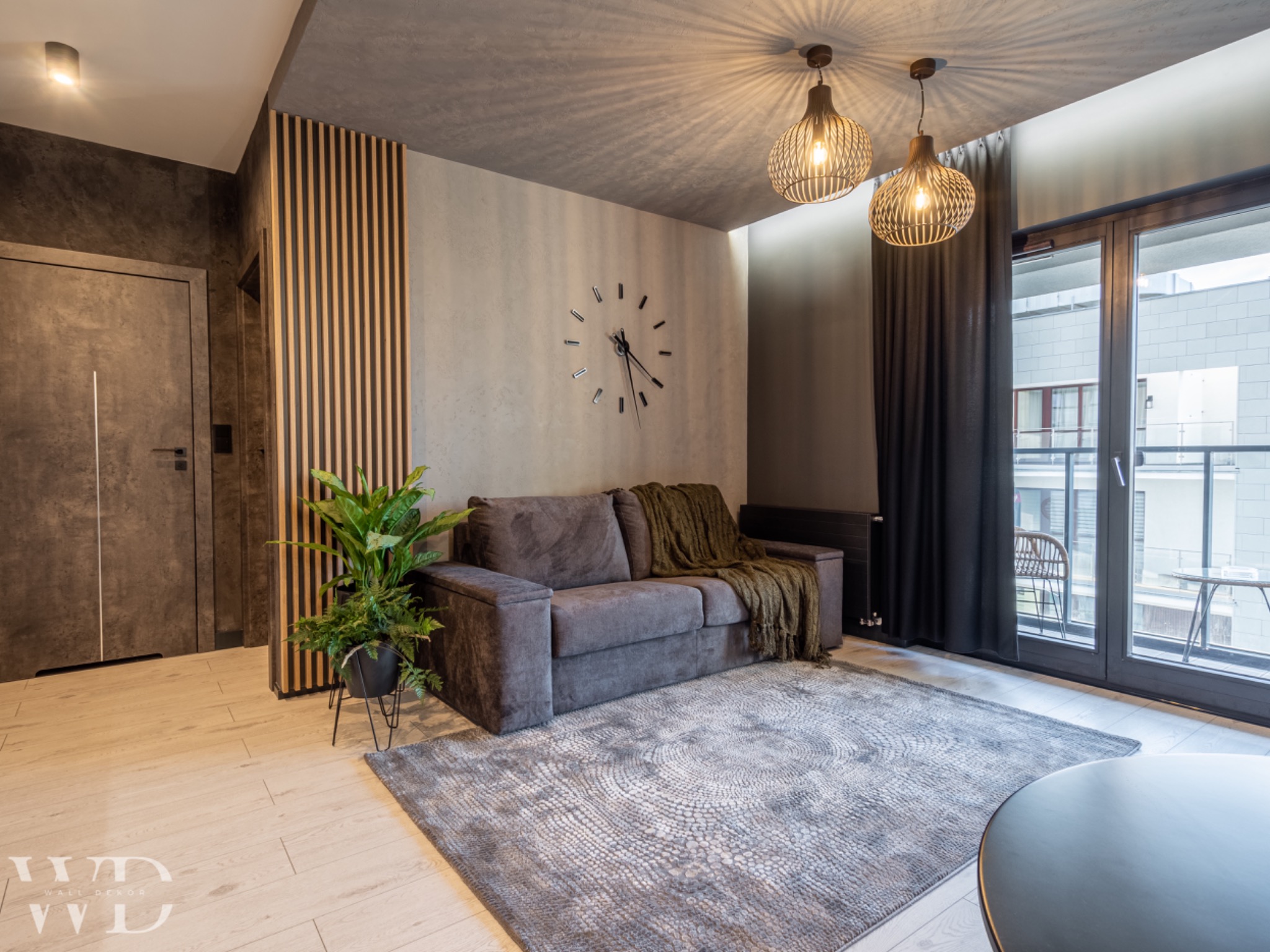 Nowoczesny salon z szarą sofą, dywanem w geometryczne wzory, drewnianymi lamelami na ścianie oraz lampami wiszącymi z ażurowymi kloszami, widok na balkon.