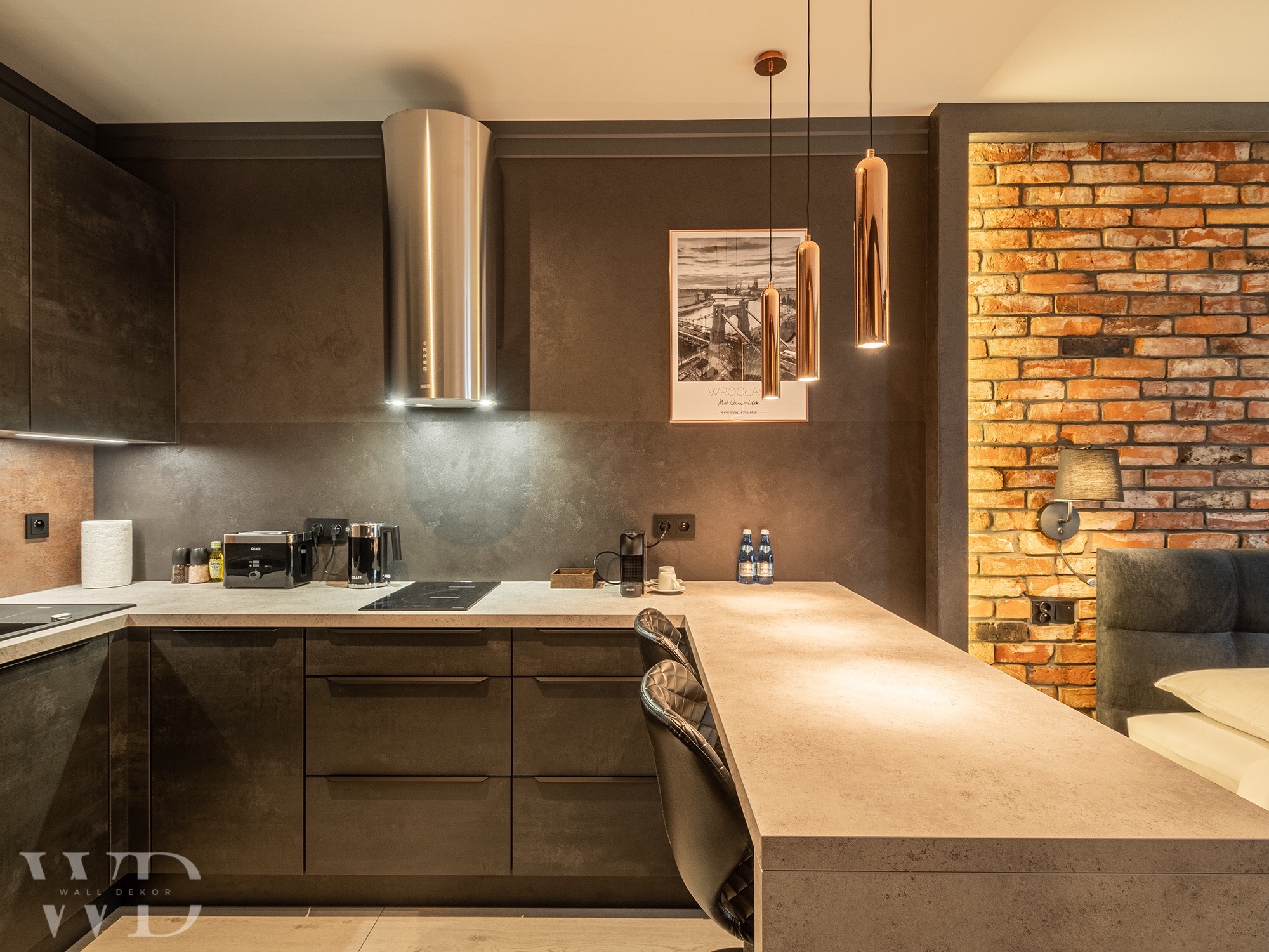 Nowoczesna kuchnia z ciemnymi, matowymi szafkami, betonowym blatem i ceglaną ścianą w tle, oświetlona miedzianymi lampami wiszącymi i srebrnym okapem.