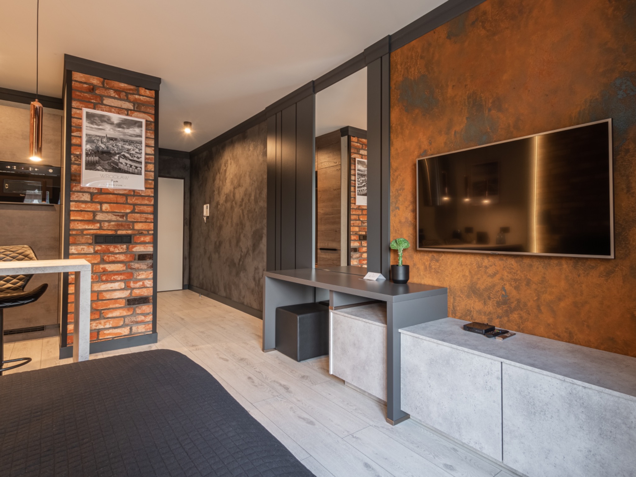 Nowoczesne wykończenie wnętrza mieszkania z akcentami cegły i tynku dekoracyjnego w odcieniach brązu, szarości i bieli, widoczny telewizor i meble w minimalistycznym stylu.