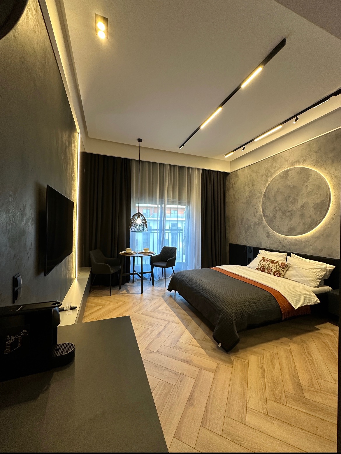 Nowoczesna sypialnia z łóżkiem, stolikiem i krzesłami, oświetleniem LED w suficie i na ścianie, oraz podłogą ułożoną w jodełkę.