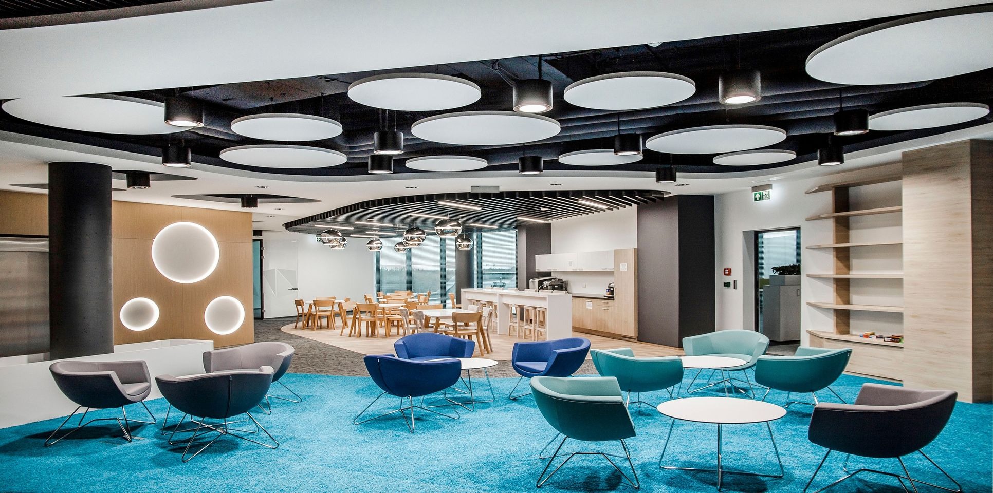 Nowoczesna aranżacja biura z niebieską wykładziną dywanową, designerskimi fotelami w odcieniach szarości, błękitu i turkusu, okrągłymi stolikami oraz strefą jadalną w tle.