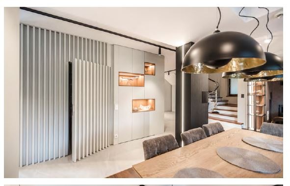 Nowoczesne wnętrze jadalni z drewnianym stołem, szarymi krzesłami, wiszącymi lampami z elementami złota i zabudową meblową z podświetlanymi niszami ekspozycyjnymi oraz ścianą z dekoracyjnych...