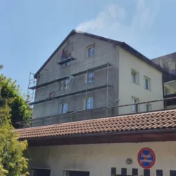 Pracownik na rusztowaniu myje elewację budynku strumieniem wody pod ciśnieniem, widoczne zacieki na ścianie i fragment dachu garażu z oznaczeniami parkingu.