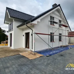 Nowoczesny dom jednorodzinny z białą elewacją, w trakcie prac wykończeniowych; widoczne rusztowania, zabezpieczona folią podstawa, czarny dach z dachówki ceramicznej, szara kostka brukowa...