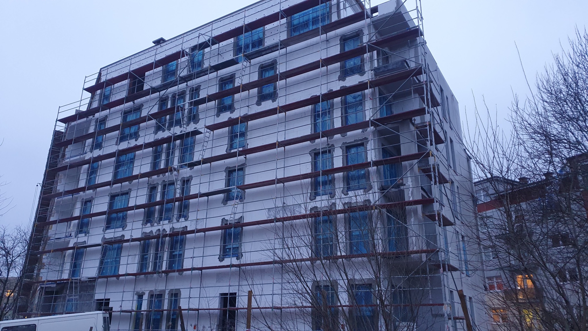 Wielopiętrowy budynek w trakcie ocieplania, osłonięte folią okna, rusztowania na całej elewacji, widoczne ślady kleju na styropianie, pochmurne niebo w tle.