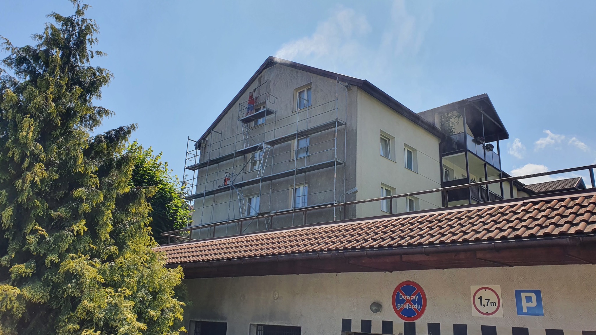 Pracownik na rusztowaniu myje elewację budynku strumieniem wody pod ciśnieniem, widoczne zacieki na ścianie i fragment dachu garażu z oznaczeniami parkingu.