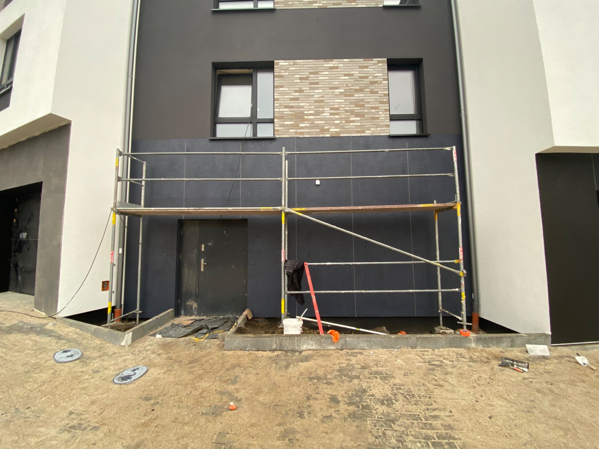 Dwukondygnacyjny budynek z ciemnoszarą elewacją, częściowo pokrytą rusztowaniem, z fragmentem elewacji wykończonym cegłą klinkierową, widoczne narzędzia i materiały budowlane.