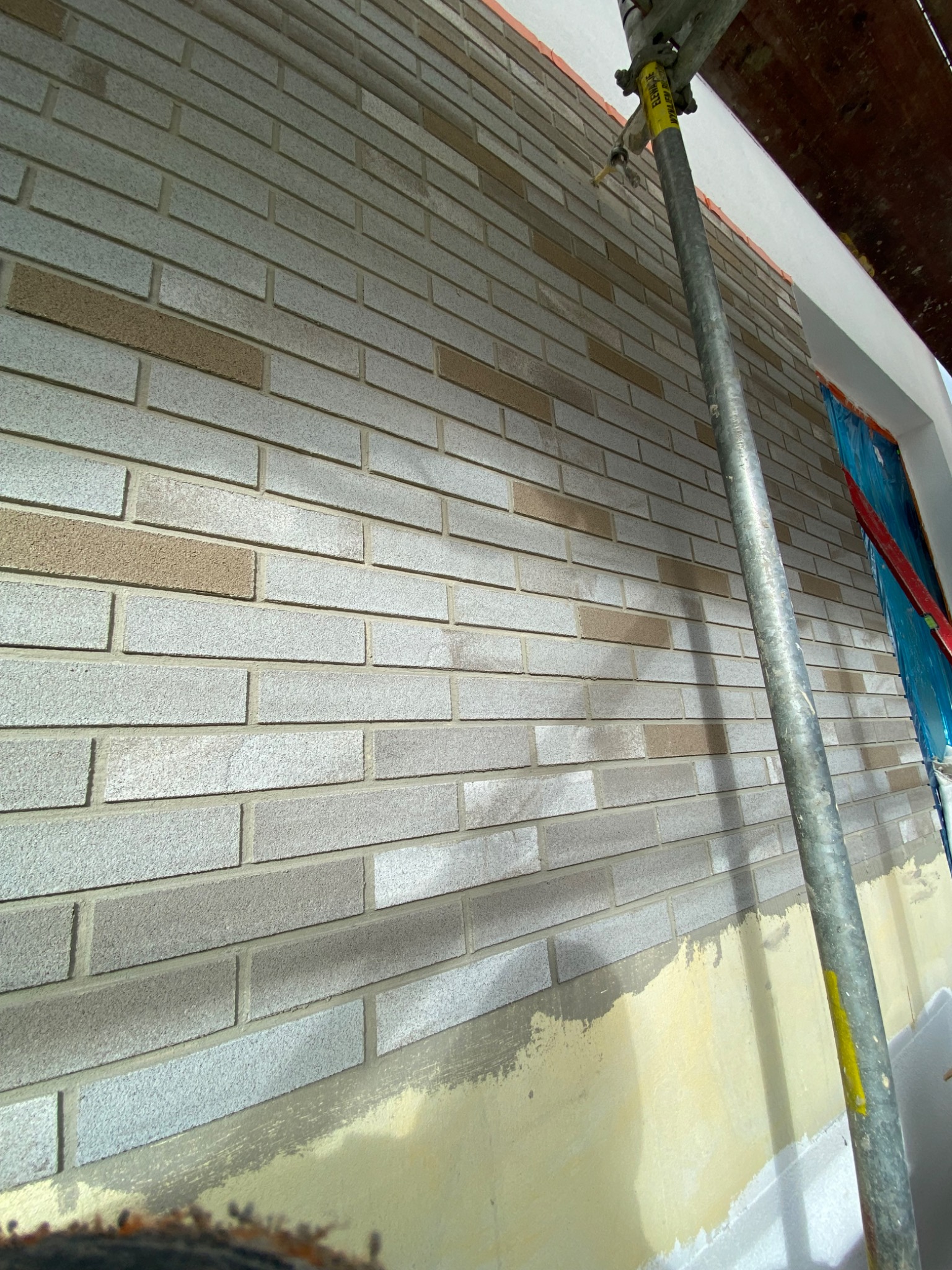 Świeżo położona elewacja z szarej cegły klinkierowej z akcentami beżowymi, widoczny fragment rusztowania i zabezpieczone okno folią malarską, stan surowy na dole ściany.