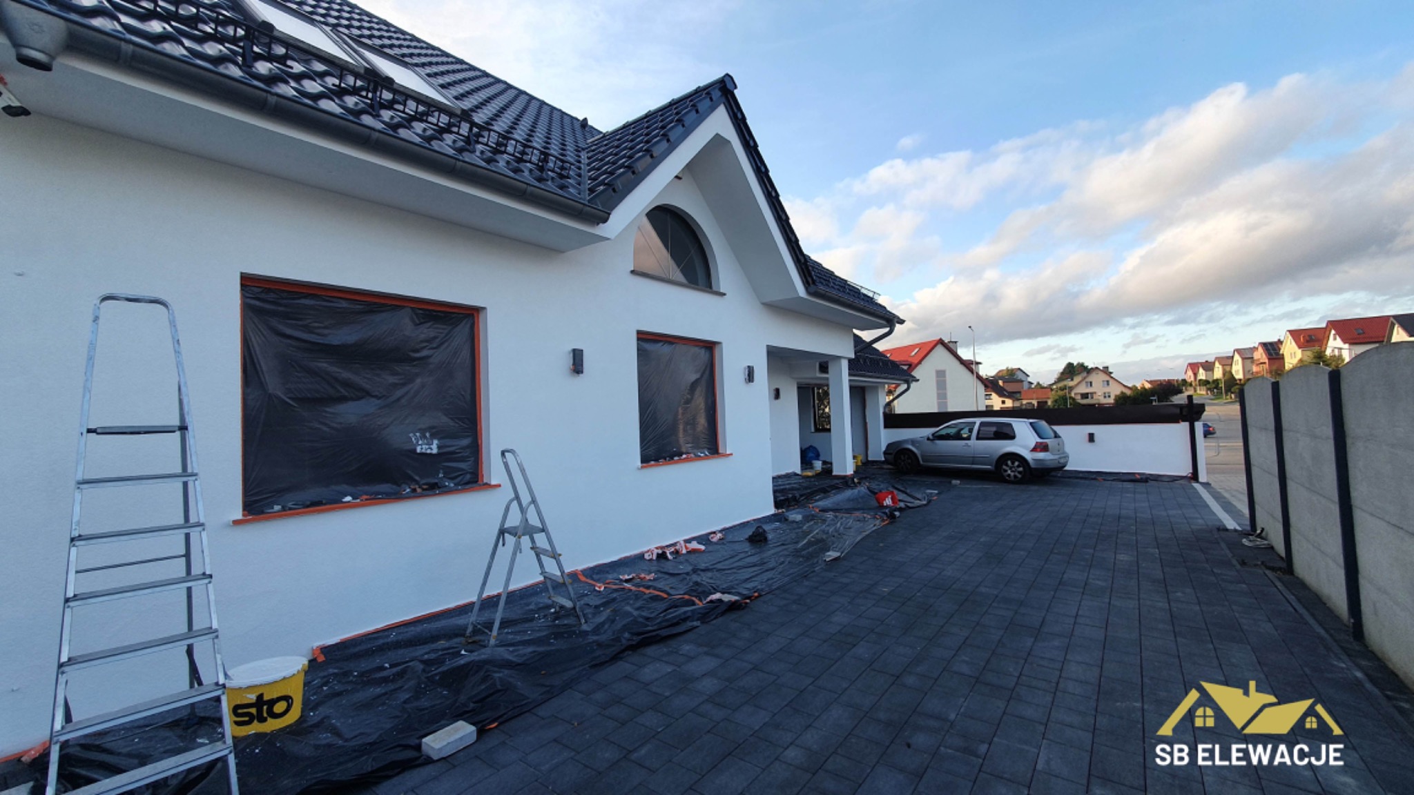 Biały dom jednorodzinny z czarną dachówką w trakcie prac elewacyjnych, okna zabezpieczone folią, widoczne drabiny i wiadro farby Sto, teren wokół zabezpieczony folią malarską.