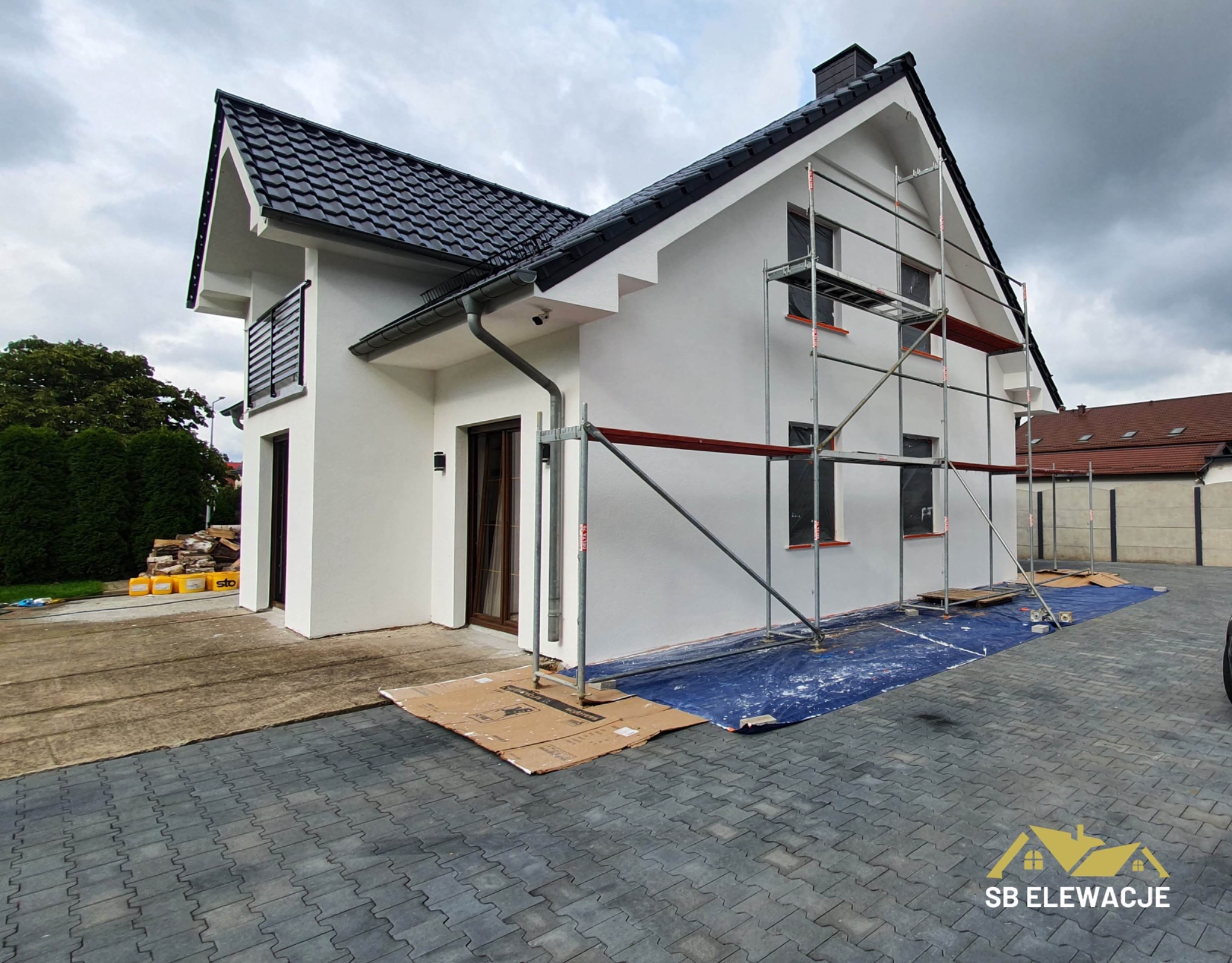 Nowoczesny dom jednorodzinny z białą elewacją, w trakcie prac wykończeniowych; widoczne rusztowania, zabezpieczona folią podstawa, czarny dach z dachówki ceramicznej, szara kostka brukowa...