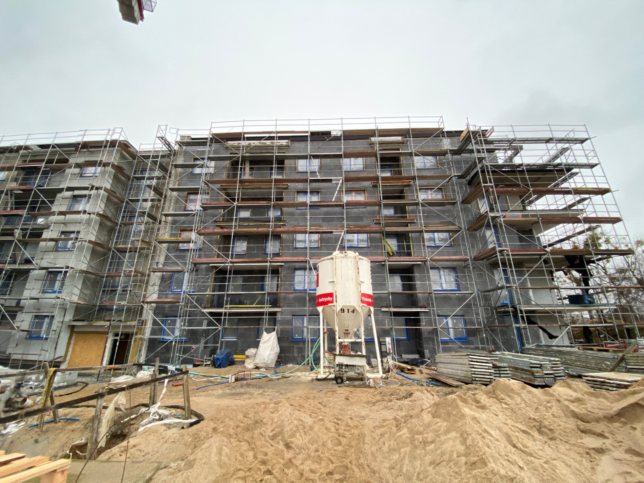 Budynek wielorodzinny w Trzebielinie w trakcie ocieplania, obłożony rusztowaniami, z widoczną maszyną do podawania zaprawy i stertą piasku na pierwszym planie.