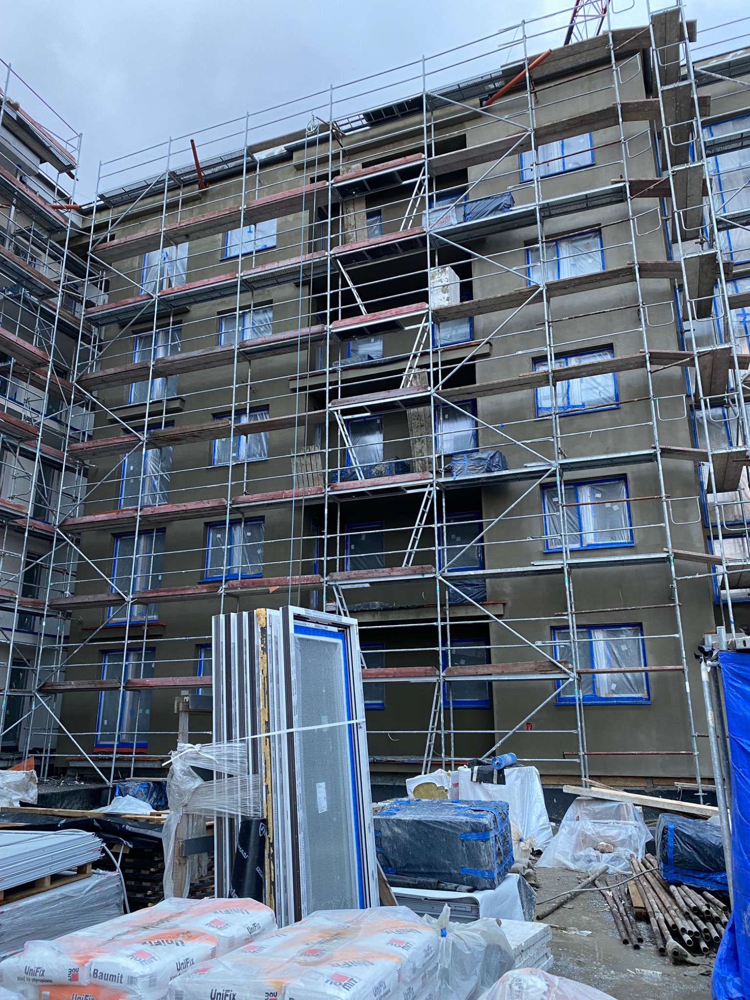 Budynek wielorodzinny w trakcie prac elewacyjnych: rusztowania, zabezpieczone okna folią, przygotowane worki z zaprawą i nowe okna.