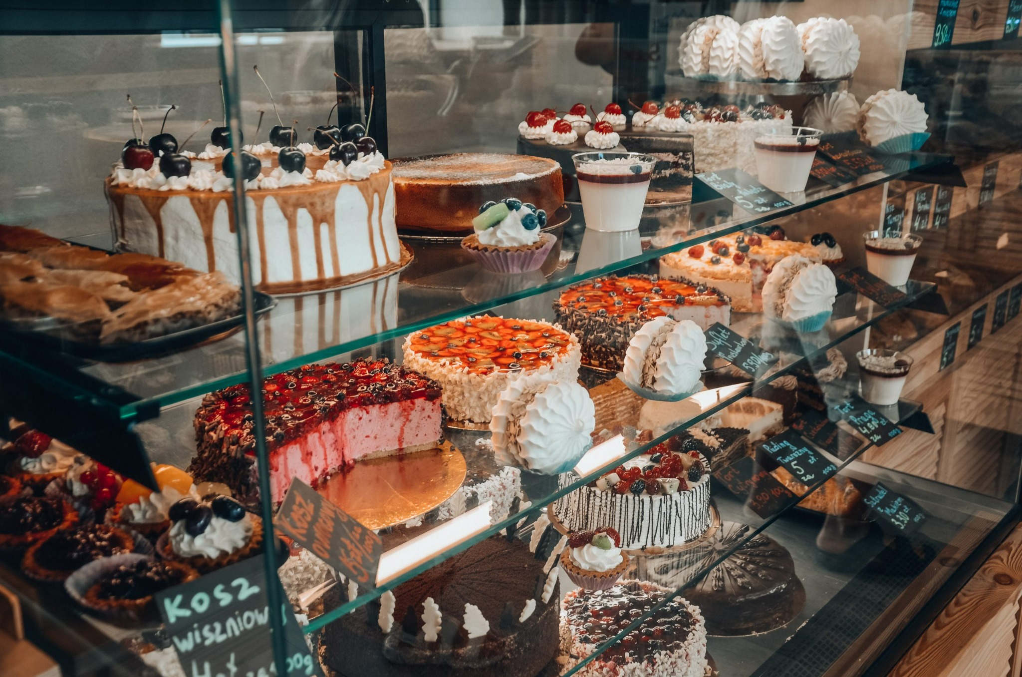 Słodki asortyment w witrynie: tort z wiśniami i polewą karmelową, sernik, babeczki z owocami, deser w szklance, tort truskawkowy, bezy oraz tabliczki z cenami.
