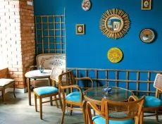 Jasne wnętrze kawiarni z niebieską ścianą ozdobioną lustrami i obrazkami, drewnianymi krzesłami z turkusowymi poduszkami i stolikami. Fragment ściany z cegły i bambusowe elementy dekoracyjne.