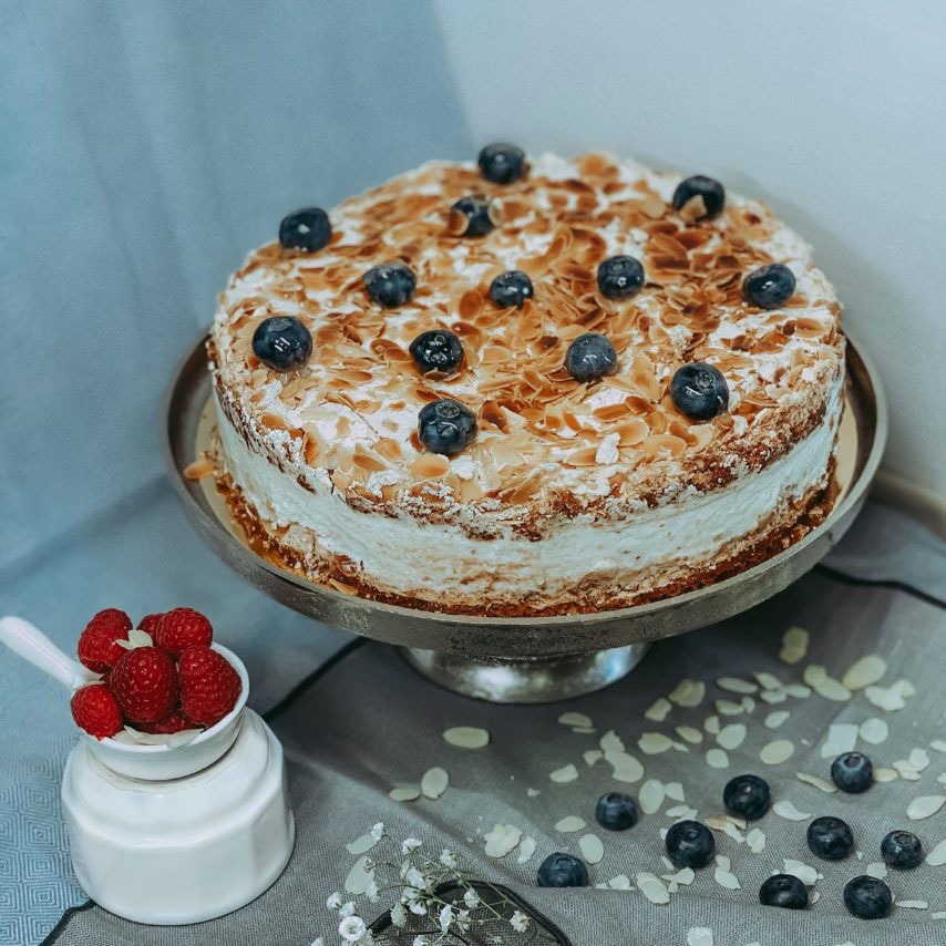 Tort bezowy z płatkami migdałów i borówkami, podany na srebrnej paterze, obok biały pojemnik z malinami, dekoracja z drobnych kwiatów na szarym tle