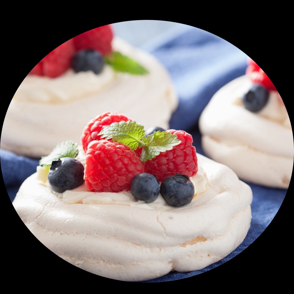 Trzy mini bezy Pavlova z kremem, malinami, borówkami i miętą na niebieskiej tkaninie, widoczne z bliska.