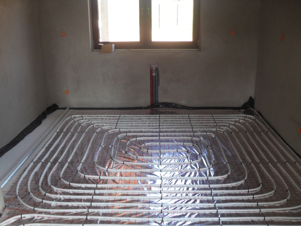 Instalacja ogrzewania podłogowego: białe rury ułożone spiralnie na folii odblaskowej, przymocowane do czerwonej siatki w surowym wnętrzu z oknem.
