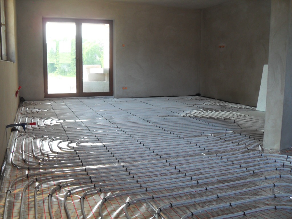 Montaż ogrzewania podłogowego: rury grzewcze ułożone na folii izolacyjnej w surowym wnętrzu z oknem balkonowym w tle.