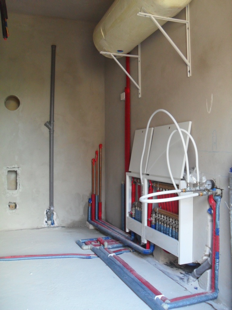 Rozdzielacz ogrzewania podłogowego z licznymi rurami w nowym budynku, zbiornik wyrównawczy na ścianie, surowy stan wykończenia, widoczne instalacje w podłodze i na ścianach.