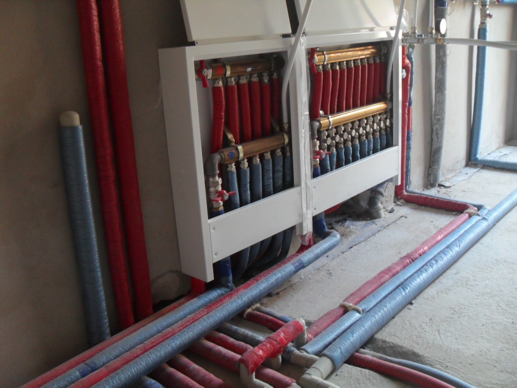 Rozdzielacz instalacji grzewczej z rurami w izolacji czerwonej i szarej, widoczne zawory i przyłącza, surowy stan wykończenia pomieszczenia.
