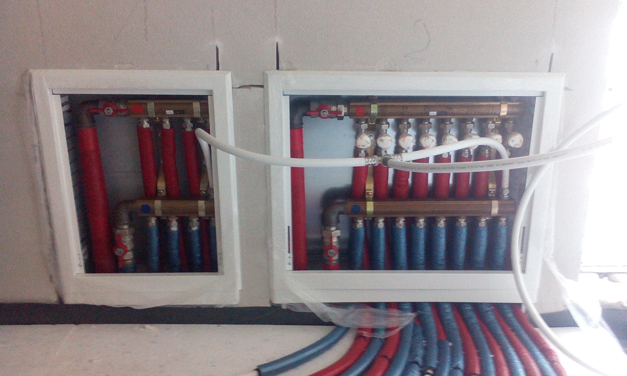Rozdzielacz instalacji podłogowej w dwóch białych szafkach, z widocznymi czerwonymi i niebieskimi rurami oraz zaworami regulacyjnymi, częściowo zasłonięty folią ochronną.