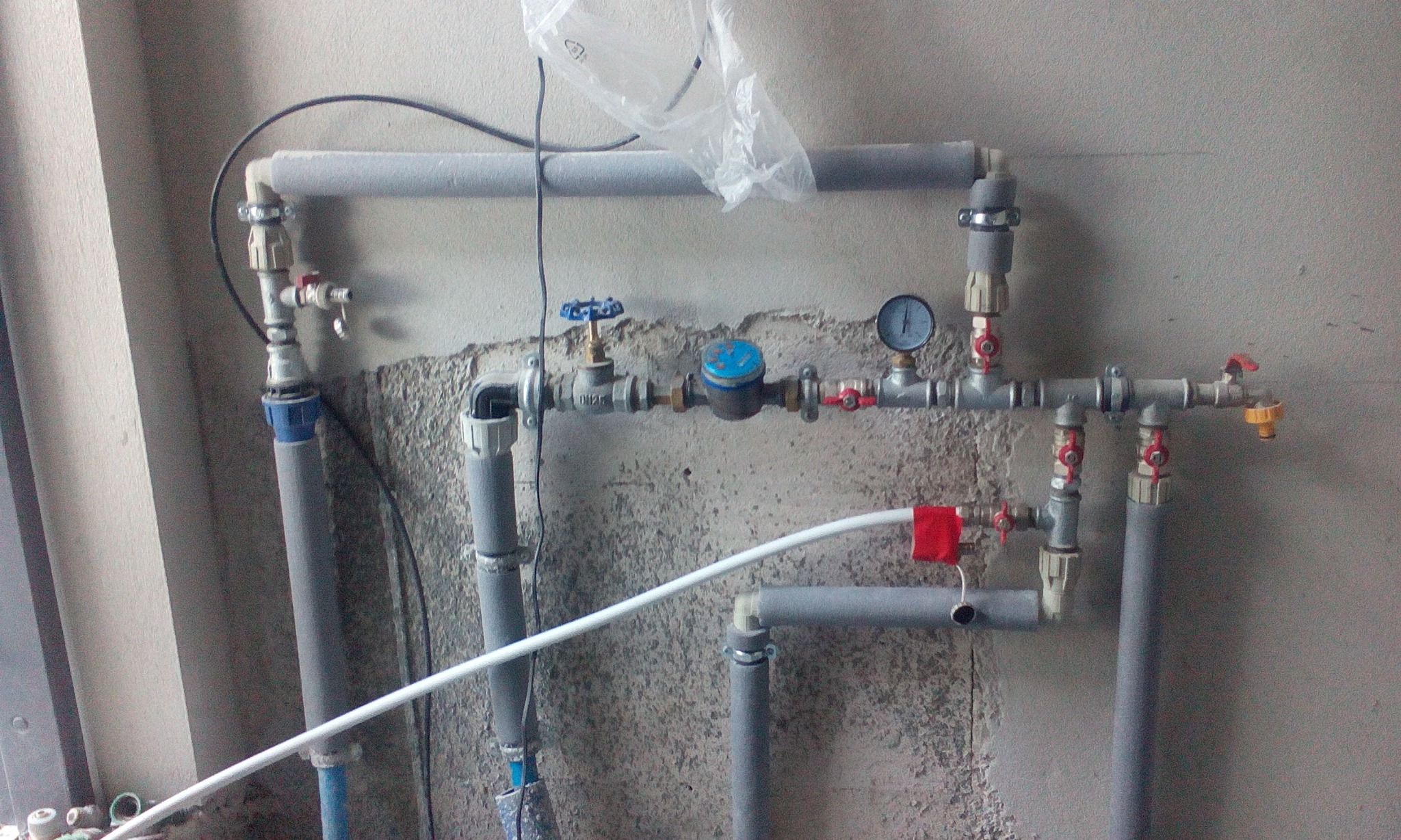 Szare rury instalacji wodnej z zaworami, manometrem i licznikiem na tle surowej ściany z widocznym tynkiem.