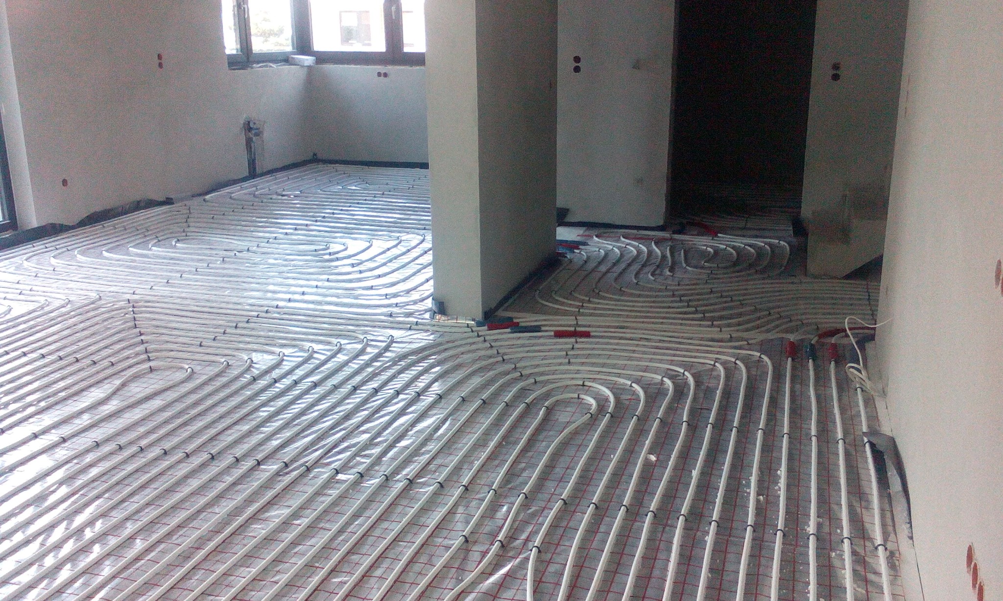 Białe rury ogrzewania podłogowego ułożone na siatce w surowym wnętrzu, widok z góry, w tle filary i okna.