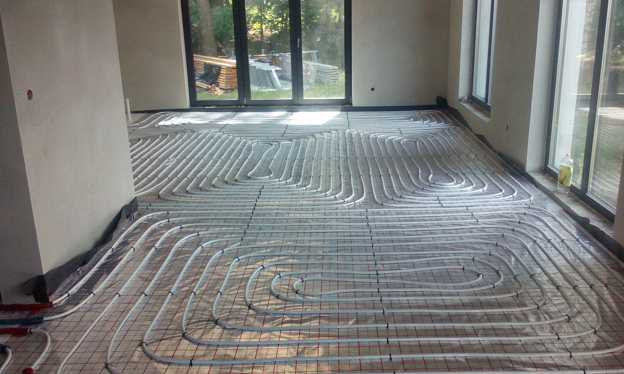 Rozłożone rury ogrzewania podłogowego na siatce w surowym wnętrzu, widoczne okna i materiały budowlane w tle.