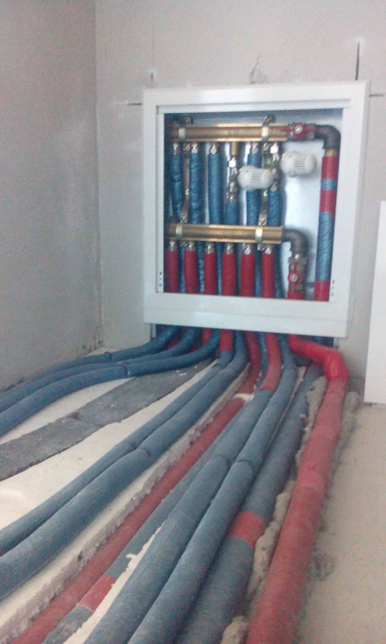 Rozdzielacz instalacji centralnego ogrzewania w szafce, z widocznymi rurami doprowadzającymi w kolorach czerwonym i niebieskim, ułożonymi w bruzdach w podłodze.