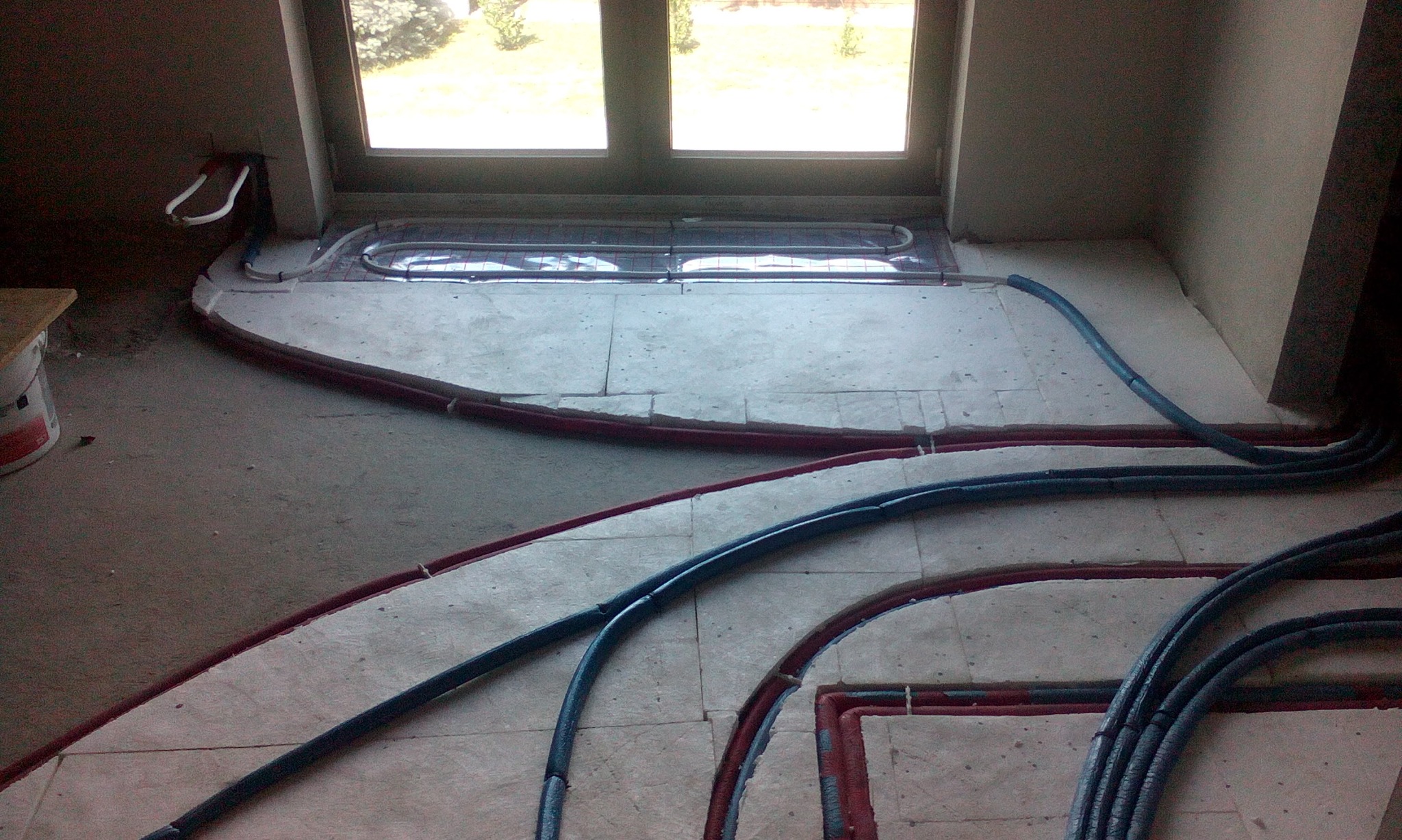 Rozłożone rury ogrzewania podłogowego w nowym budownictwie, widoczne czerwone i niebieskie przewody w trakcie montażu przed wylewką, okno w tle.