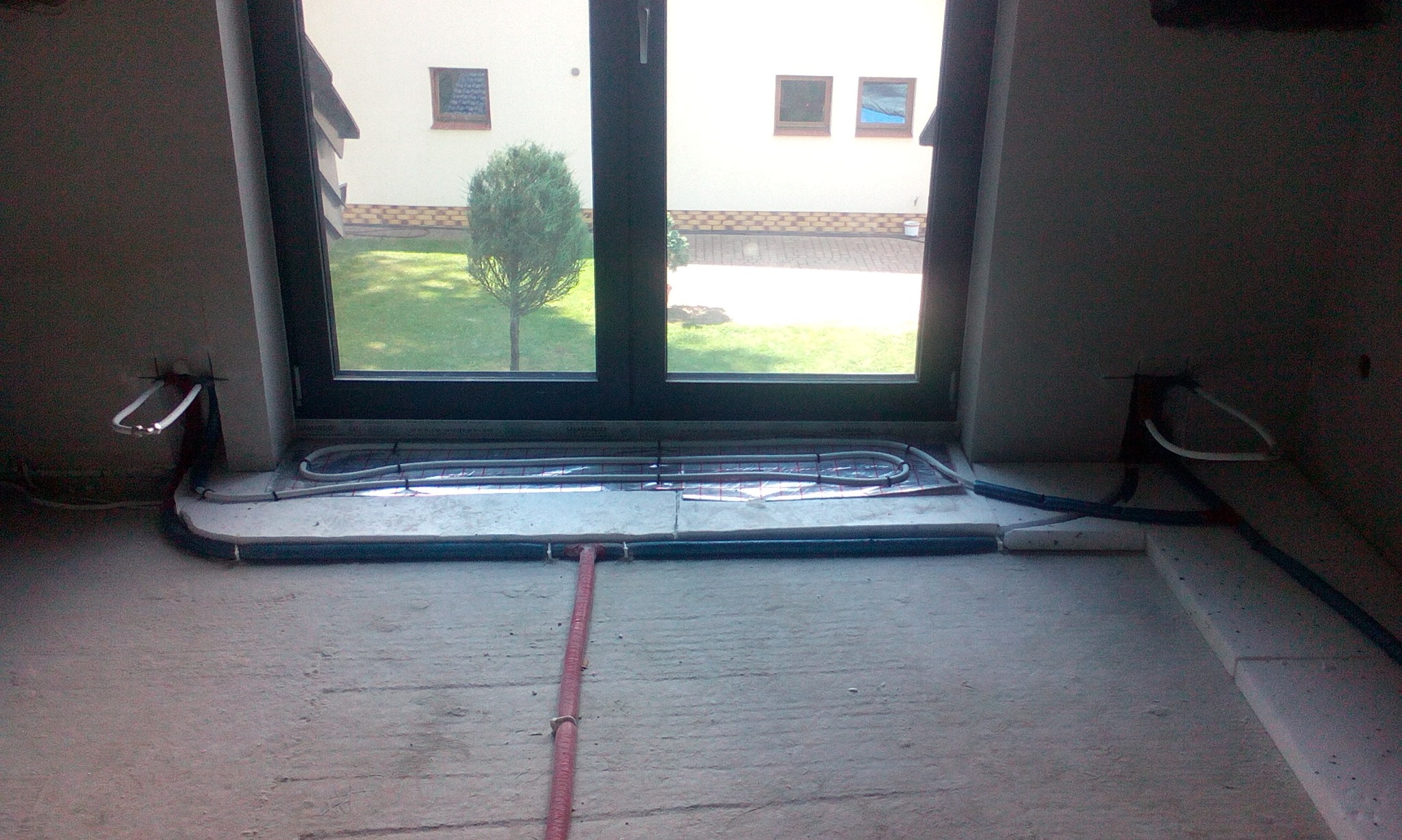 Montaż ogrzewania podłogowego przy oknie, widoczne rury grzewcze na folii izolacyjnej, fragmenty ścian w surowym stanie.