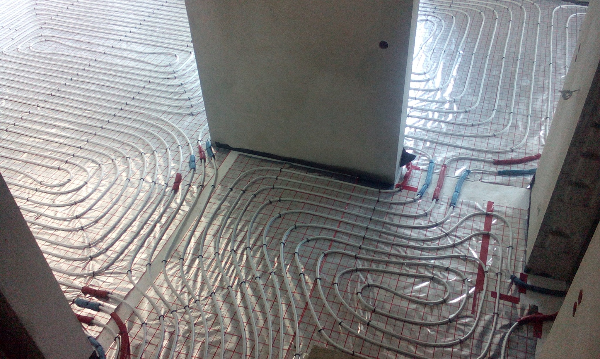 Białe rury ogrzewania podłogowego ułożone na folii z czerwoną siatką, widoczne fragmenty ścian i izolacji.