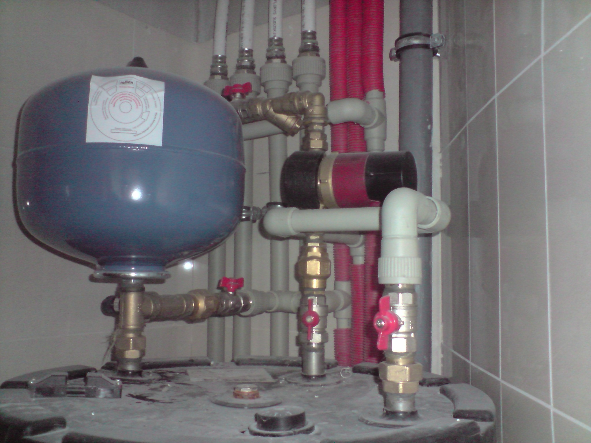 Niebieski zbiornik wyrównawczy z naklejoną instrukcją, podłączony do systemu rur z zaworami o czerwonych uchwytach w pomieszczeniu z wykafelkowanymi ścianami.
