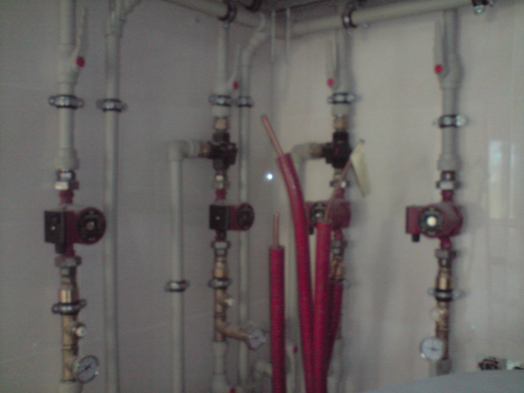 Fragment instalacji grzewczej z czterema pionami rur, pompami obiegowymi i manometrami, widoczne rury miedziane w izolacji.