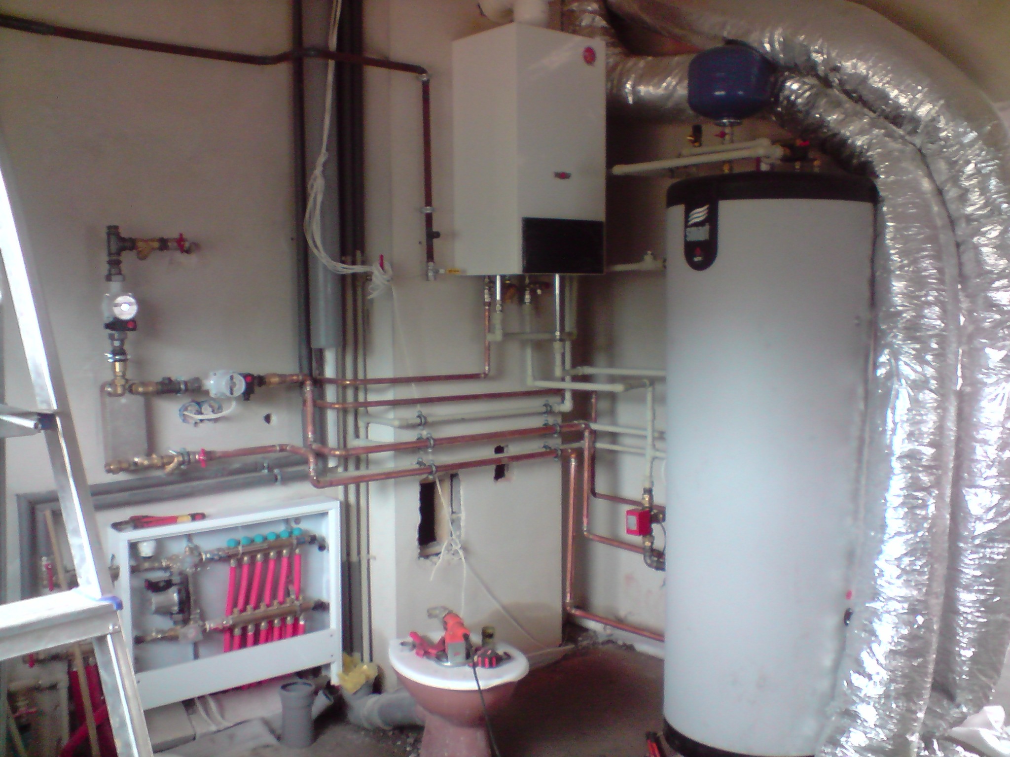 Kotłownia w trakcie instalacji: widoczny zasobnik ciepłej wody z izolacją, piec gazowy, rozdzielacz ogrzewania podłogowego z czerwonymi pętlami, rury miedziane, drabina i narzędzia na sedesie.