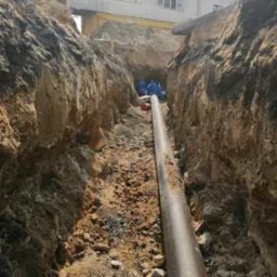 Rura ułożona w wykopie ziemnym, widoczni pracownicy w oddali, budynek w tle.