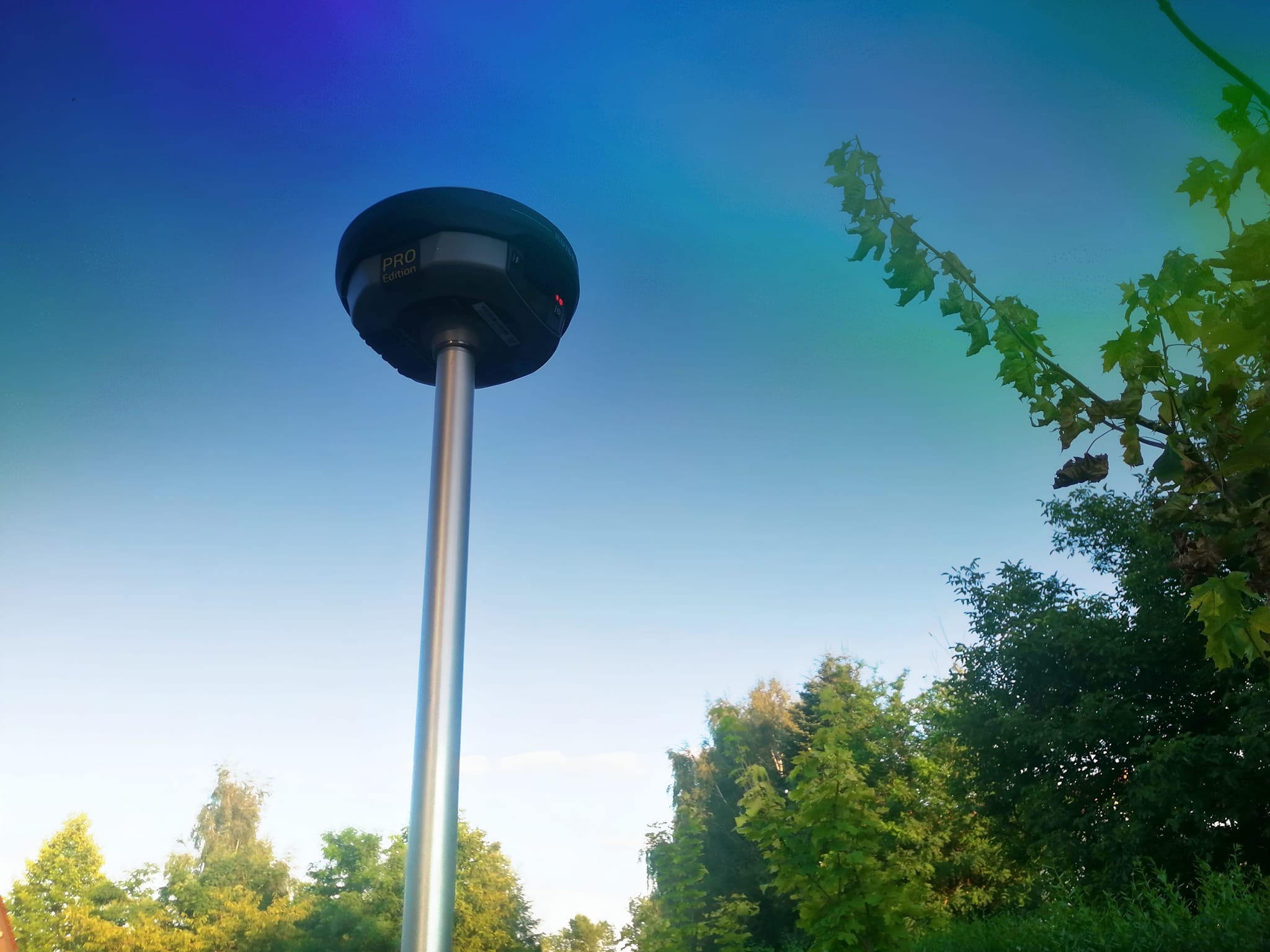 Geodezyjny odbiornik GNSS 'PRO Edition' na maszcie, skierowany w niebo z widocznymi drzewami w tle.